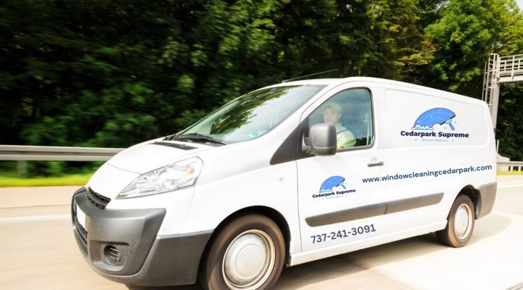 A white van traveling on the highway, featuring a window cleaning service advertisement and a visible contact number.